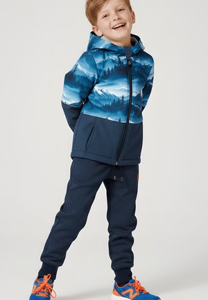 Uśmiechnięte dziecko ubrane w niebieską kurtkę z kapturem i nadrukiem gór, granatowe spodnie dresowe oraz niebieskie trampki z pomarańczowymi sznurówkami stoi z rękoma za plecami.