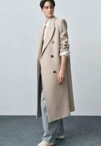 Cappotto di lana beige con chiusura doppio petto, ampi revers e bottoni neri. Indossato sopra un maglione chiaro e pantaloni grigi.