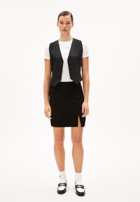 Zwarte getailleerde vest met knopen over een wit T-shirt, gecombineerd met een zwarte minirok met een zijsplit. Het model draagt zwarte loafers en witte sokken.
