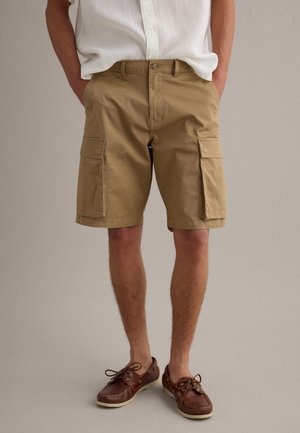 Uomo che indossa pantaloncini cargo color sabbia fino al ginocchio, camicia bianca a maniche corte e scarpe da barca in pelle marrone, in piedi con le mani nelle tasche.