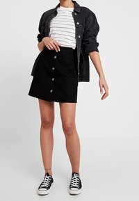 Falda de mezclilla negra con botones frontales, combinada con una camiseta a rayas y una chaqueta de mezclilla negra, completada con zapatillas negras.