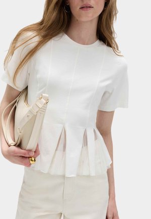 Frau trägt ein weißes, plissiertes Kurzarmoberteil und hält eine beige Handtasche mit goldfarbenen Beschlägen vor schlichtem Hintergrund.