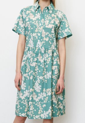 Kobieta ubrana w turkusową sukienkę z krótkim rękawem, ozdobioną białymi kwiatowymi wzorami i kołnierzykiem na guziki, stojąca na tle jednolitego białego tła.