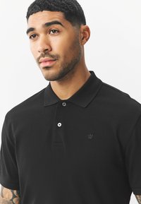 Mustast tekstuurkangast polo-särk, millel on krae ja kaks nööpi ning rinnanahal peen kroon-logo.