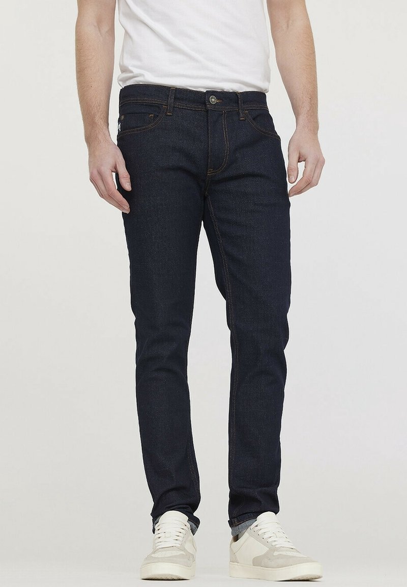 Homme portant un jean slim bleu foncé, des baskets blanches et un t-shirt blanc, debout devant un fond clair uni.