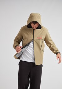 Chaqueta impermeable beige con capucha, cremallera frontal y detalles en negro; cuenta con un bolsillo interior y una etiqueta con el logo en el pecho.