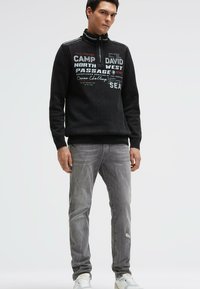 Schwarzer Strickpullover mit hohem Kragen, versehen mit weißen und roten Textdesigns; kombiniert mit hellgrauen Jeans und weißen Turnschuhen.