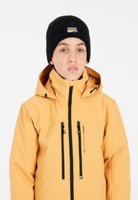 Veste imperméable jaune avec des fermetures éclair noires et deux poches avant, associée à un bonnet côtelé noir avec un patch logo.