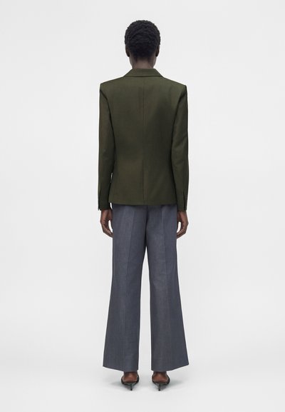 Άτομο που φοράει σακάκι σε ελαιοπράσινο χρώμα, γκρι φαρδιά παντελόνια και μαύρα τακούνια, στέκεται γυρισμένο με την πλάτη σε απλό λευκό φόντο.