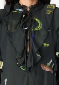 Blouse noire transparente avec un col à volants, présentant des motifs floraux multicolores et des champignons complexes, et une fermeture à nouer à l'avant.