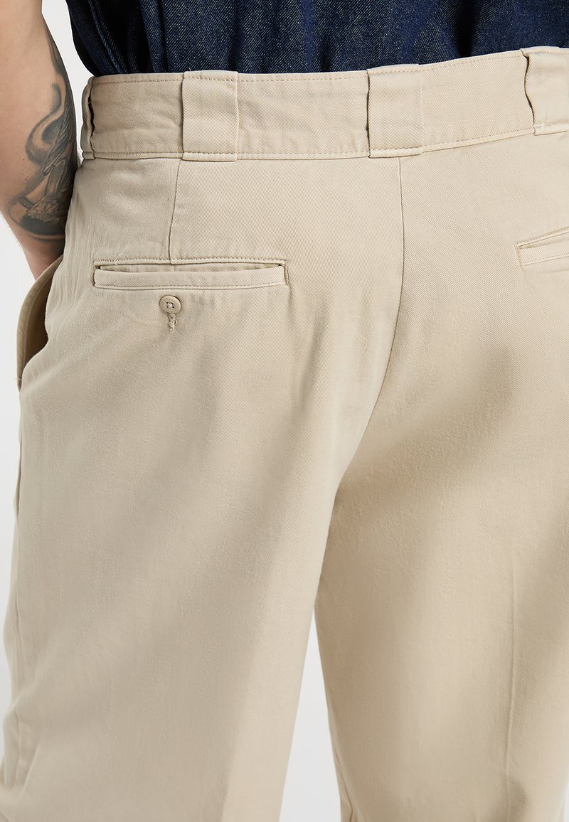 Pantaloni beige con tasca posteriore con bottone indossati da una persona con un braccio tatuato e camicia scura infilata.