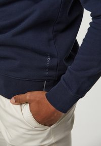 Marinblå sweatshirt med ribbade muddar och fåll, med en subtil logotyp på sidan. Mjukt bomullstyg och strömlinjeformad design. Ljusbeiga byxor.