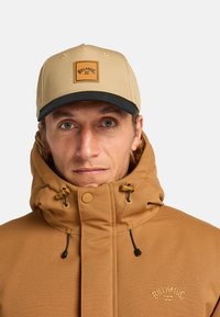 Giacca con cappuccio marrone con una superficie testurizzata e chiusure con coulisse; cappellino beige con patch logo; sfondo neutro.