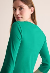 Grön långärmad topp med rundad halsringning, figursydd design och slät textur, visad bakifrån med en rak silhuett.