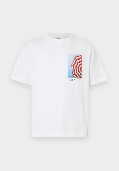 Λευκό T-shirt με φωτογραφία μιας ομπρέλας με κόκκινα και λευκά ριγέ, μπροστά σε γαλάζιο ουρανό, με το κείμενο «ΚΑΛΕΣ ΣΤΙΓΜΕΣ ΚΑΙ ΡΑΓΑΔΕΣ ΤΑΝ» από κάτω.