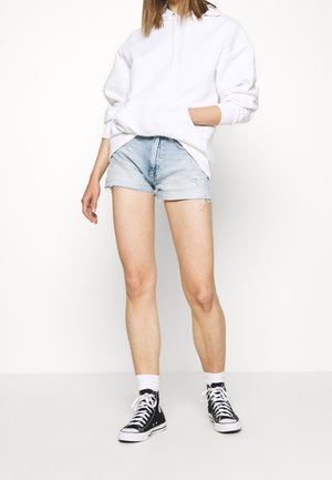 Felpa bianca con cappuccio, pantaloncini in denim azzurri con orlo sfrangiato e scarpe sneakers alte nere con suole bianche e calze nere.