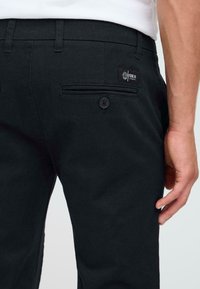 Zwarte katoenen broek met een knoopzak aan de achterkant, een klein merkembleem en een getailleerde pasvorm. De stof heeft een soepele textuur.