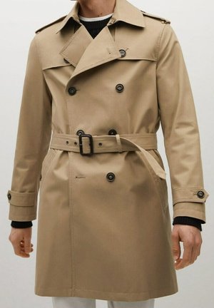 Trench-coat beige à double boutonnage avec boutons noirs, taille ceinturée et bracelets aux poignets, porté sur un pull noir et blanc.