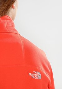 Chaqueta de forro polar roja con un cuello alto, que presenta un logo bordado en la parte posterior. La tela parece suave con una textura ligera.