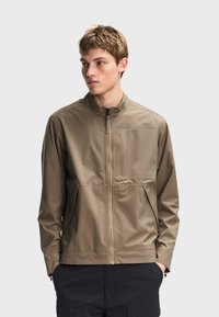 Νέος άνδρας που φοράει ανοιχτό καφέ μπουφάν με φερμουάρ, υψηλό γιακά και τσέπες, στέκεται με τα χέρια στις τσέπες του σκούρου παντελονιού, κοιτάζοντας προς τα αριστερά.