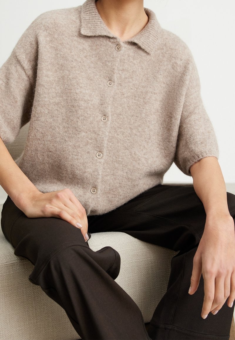 Pull en maille beige à manches courtes avec un col côtelé et six boutons, accompagné d'un pantalon marron foncé. Placés sur une surface claire texturée.