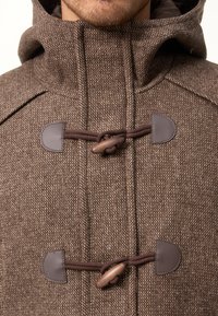 Brun ullrock med texturerad yta, hög krage och läderknäppen. Inkluderar en huva och detaljer med kontrastsömmar.