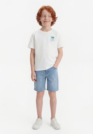 Rødhåret dreng står med hånden i lommen iført hvid t-shirt med palmetræsgrafik, denimshorts og hvide sneakers foran hvid baggrund.