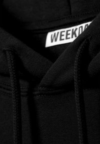 Svart hettegenser i en bomullsblanding, med snor, myk tekstur og et merke som leser "WEEKDAY" nær halsen.