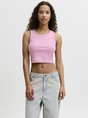 Mujer con cabello rizado que lleva un top corto rosa sin mangas y vaqueros de talle alto azul claro sobre un fondo claro y liso.