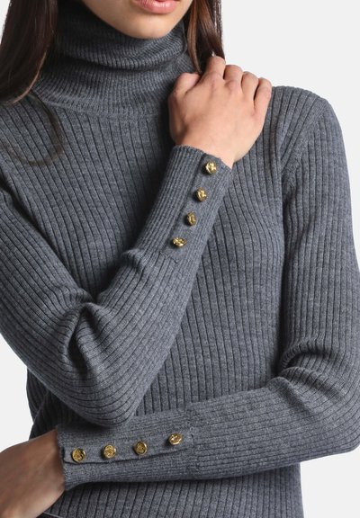 Femme portant un pull gris à col roulé côtelé avec des boutons décoratifs dorés sur les manches, bras croisés et une main sur l'épaule.