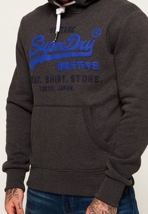 Ciemnoszara bluza z kapturem z niebieskim logo „SuperDry”, dodatkowym tekstem, białymi sznurkami i przednią kieszenią, noszona przez osobę z tatuażami.