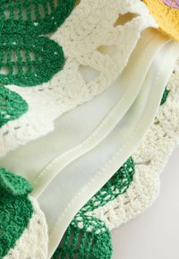 Zbliżenie na biały materiał z kremową falistą koronką na brzegu i zielonymi, szydełkowymi liśćmi na fakturowanym tkaninie.