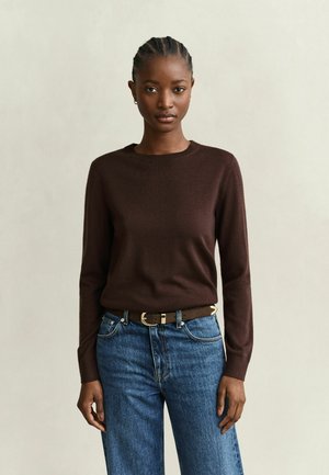 Jeune femme aux cheveux tressés, portant un pull à manches longues marron foncé, un jean bleu et une ceinture marron, debout devant un fond uni.