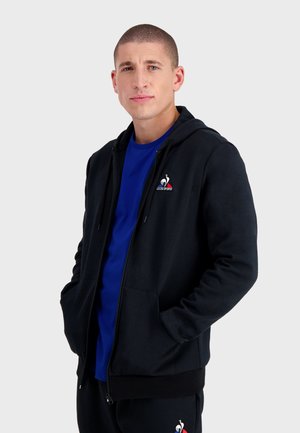 Sudadera negra con cremallera y bolsillos tipo canguro, con un pequeño logo rojo y blanco en el pecho. Lleva puesta una camiseta azul debajo. Material suave.