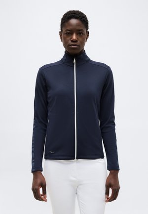 Žena má na sebe námornícky modrú bundu na zips s vysokým golierom a biele nohavice na jednoduchom svetlom pozadí.