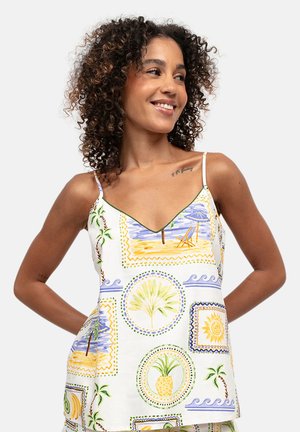 Donna con capelli ricci sorride indossando una canotta bianca con stampe tropicali di ananas, palme, sole e scene di spiaggia.