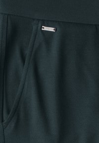 I jogger in tessuto verde scuro presentano una texture liscia, tasca laterale e un'etichetta metallica del marchio attaccata alla vita.