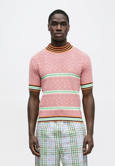 Πλεκτό πουλόβερ με κοντά μανίκια σε ροζ χρώμα με υφασμένο μοτίβο, που διαθέτει πράσινες και πορτοκαλί ρίγες στο γιακά και τα μανίκια. Πολύχρωμα καρό σορτς.