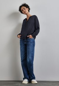 Marinebluse mit V-Ausschnitt und langen Ärmeln, kombiniert mit hellblauen Jeans und beigen Turnschuhen mit blauen Akzenten auf einem grauen Hintergrund.