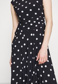 Svart klänning med vita prickar, figurnära byst och en flödande kjol. Tyget verkar slätt med en lätt lyster som framhäver mönstret.