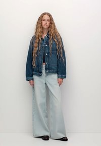 Denim jakna z udobnim krojem, svetlo modri flared jeans in črne čevlje. Dolgi, valoviti lasje obdajajo obraz, na enobarvnem ozadju.