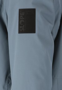 Blå vandtæt jakke med mat finish, der har et struktureret sort logo-patch mærket "SLOPE STYLE" på den øverste ærme.
