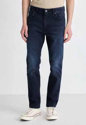 Donkerblauwe spijkerbroek met een rechte pijp, vijf zakken en een knoopsluiting. G gedragen met crèmekleurige sneakers, met witte veters.