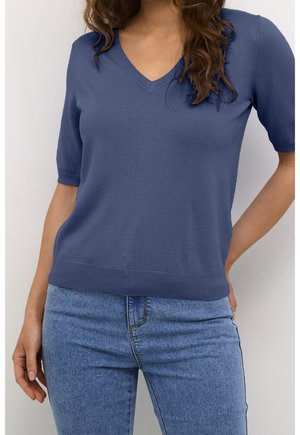 Femme portant un pull bleu à manches courtes en V et un jean en denim bleu clair, debout avec une main sur la hanche.