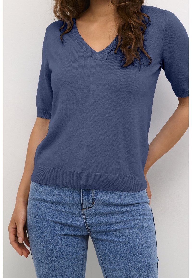 Femme portant un pull bleu à manches courtes en V et un jean en denim bleu clair, debout avec une main sur la hanche.