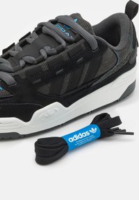 Svart och grå Adidas-sneaker med blå logofärger, vit sula och ett separat par svarta skosnören insvepta i blå Adidas-märkning.