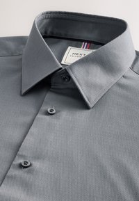 Szürke texturált ing, gombokkal, hegyes gallérral, látható márkajelzéssel és háromszínű csíkozással a nyaknál.