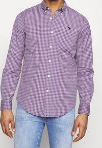 Lila- und pinkfarbener karierter Button-Up-Hemd mit langen Ärmeln, Brusttasche und weißen Knöpfen. Strukturstoff, abgerundeter Saum. Getragen mit hellblauen Jeans.