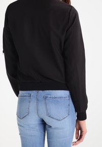 Black bomber jacket with ribbed cuffs and hem, featuring a smooth texture. Paired with light blue jeans with back pocket details.