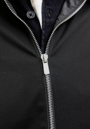 Close-up van een zilveren ritssluiting op een zwarte jas met een donker overhemd en een wit ondershirt dat eronder zichtbaar is.